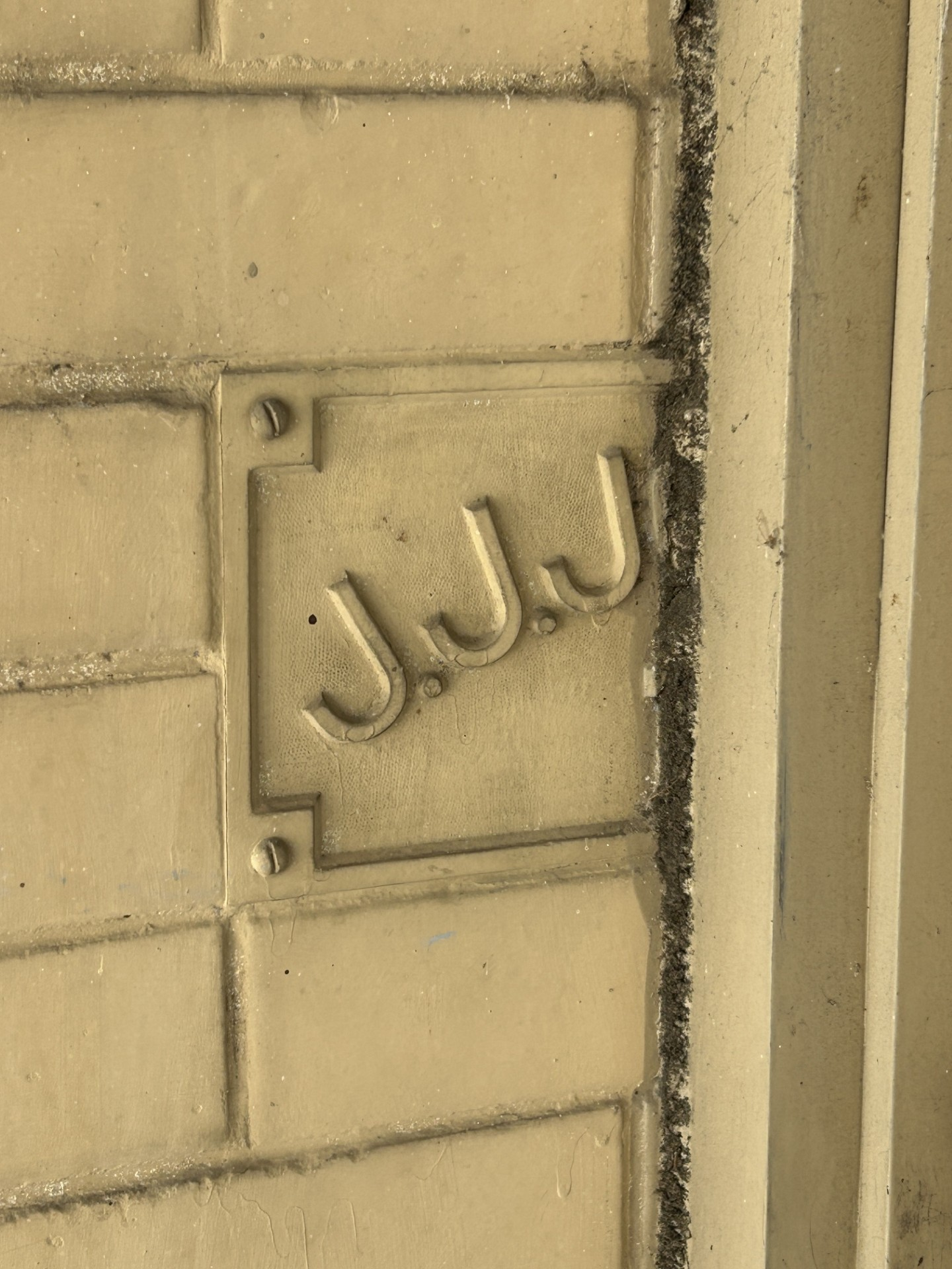 Partially hidden cast-metal plaque beside the entrance of 117 N. Main Street in downtown Harrison, showing the letters “J.J.J.” and part of what may be an “O” beneath the door frame.